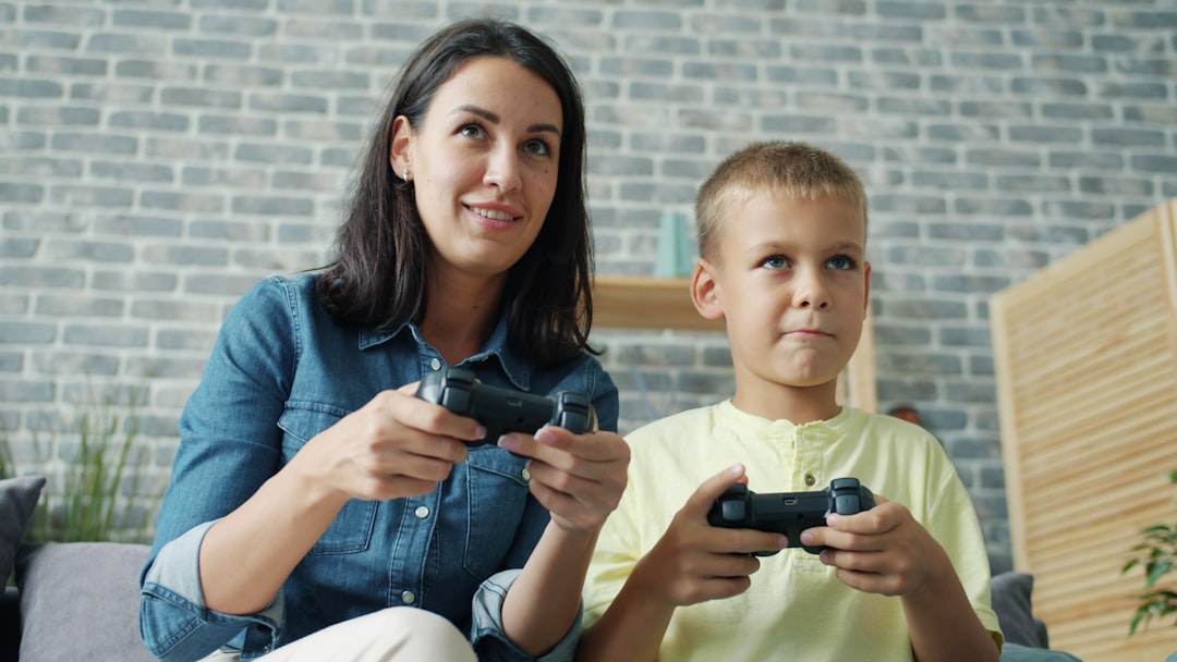 ソニーAI特許がゲーム体験を革新？自動プレイ支援の光と影 - Little boy is playing video games with caring mom holding joysticks pushing buttons and talking smiling at home. Modern devices and happy youth concept.