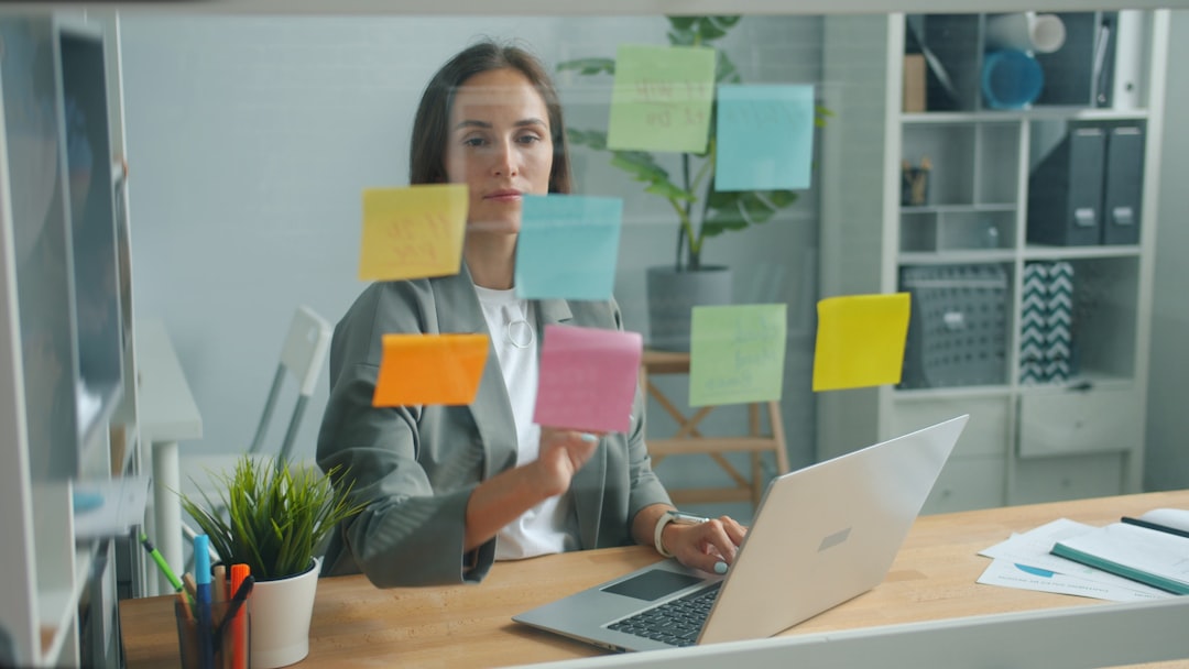 Claudeが変えるビジネスの常識：インタラクティブ分析とOffice連携の衝撃 - Good-looking young woman is working in office room using laptop behind glassboard covered with colorful stickers then taking note and reading it. Information and job concept.
