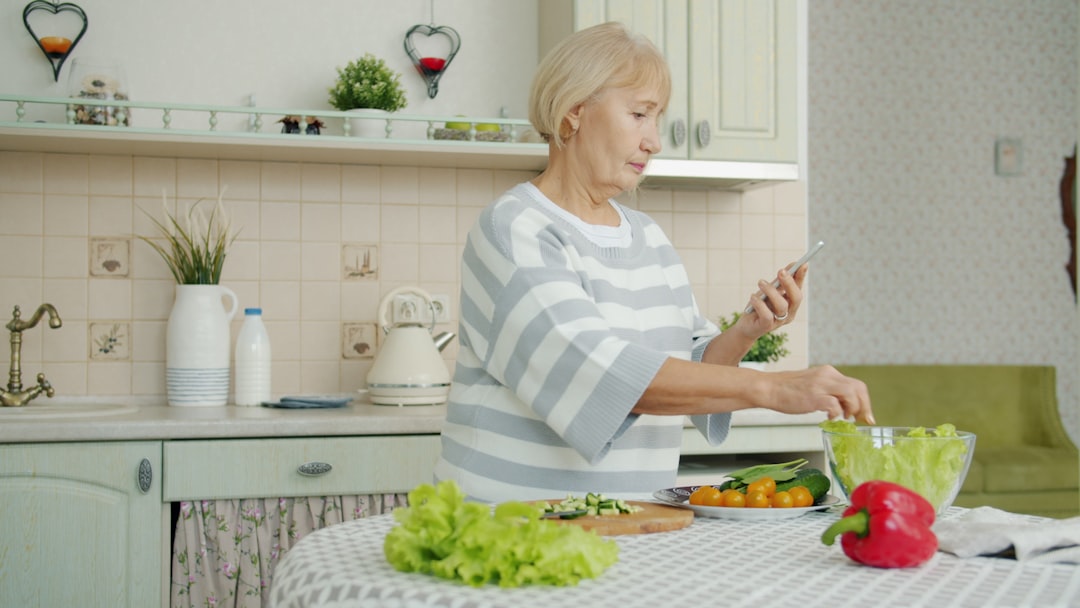 ChatGPTで再現する格安アンチエイジング術：月額20ドルの現実的アプローチ - Old lady is cooking vegetable salad and looking at smartphone screen checking ingredients for meal during dinner preparation at home. People and gadgets concept.