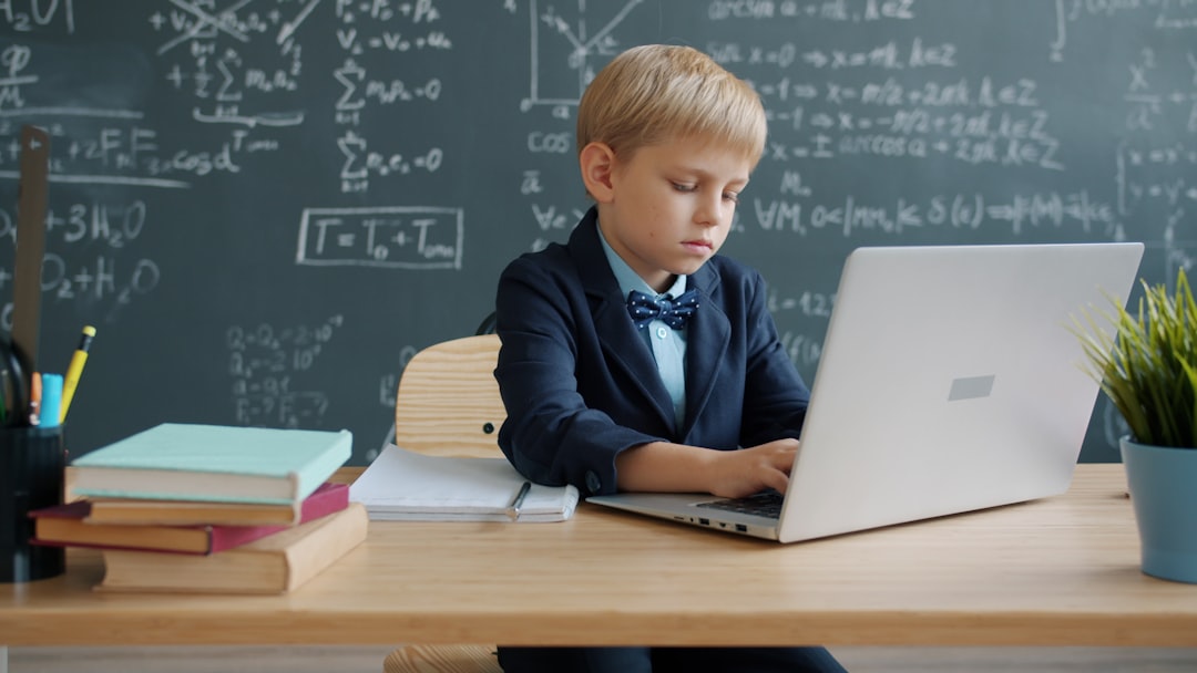 【ChatGPT】教育特化型プラグインがリリース！学習と指導を効率化 - smart little kid using laptop typing then writing at desk in university classroom, blackboard with formulas is visible in background.